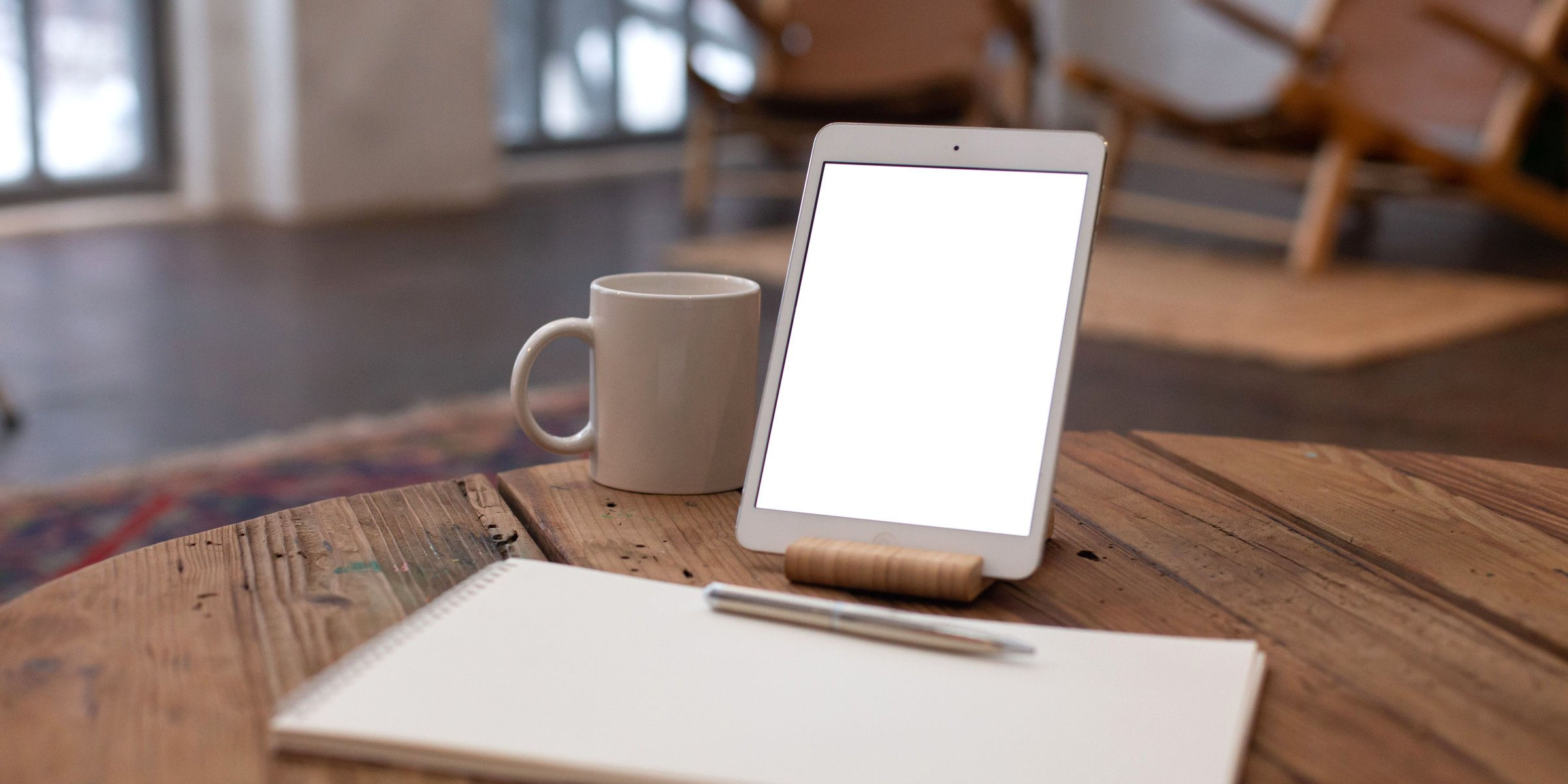 Tablet on a wood stand beside a coffee mug and notebook on a rustic wood table